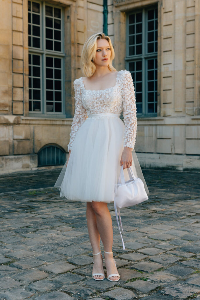 Femme qui porte une robe de mariée courte en tulle à manches longues et qui tient un sac à main.