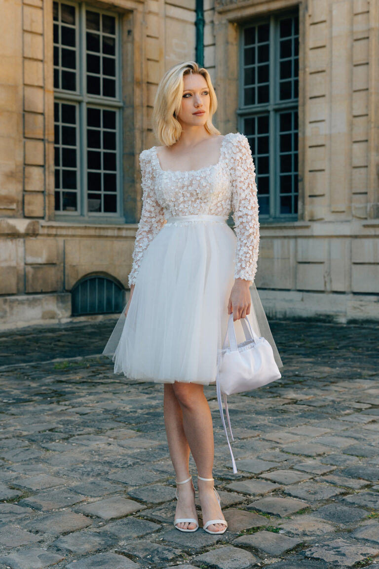 Femme qui porte une robe de mariée courte en tulle à manches longues et qui tient un sac à main.