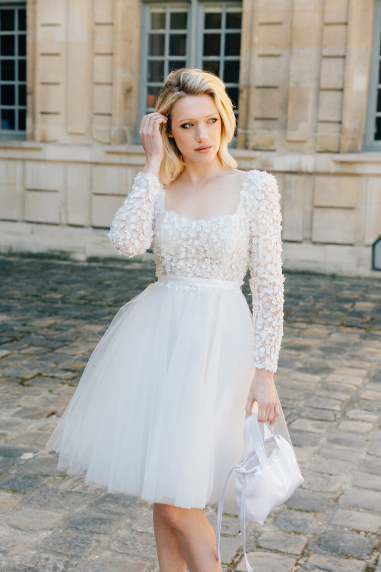 Femme qui porte une robe de mariée courte en tulle à manches longues et qui tient un sac à main.