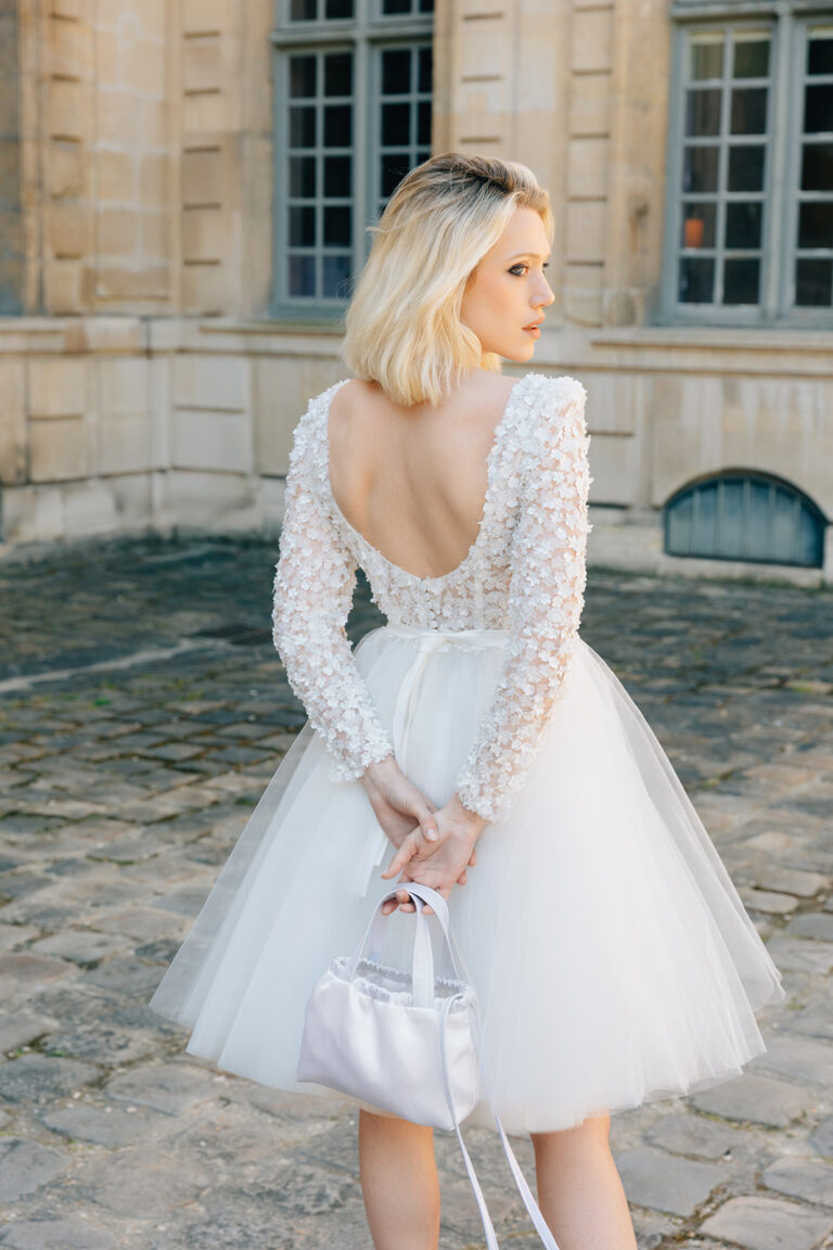 Femme de dos qui porte une robe de mariée courte en tulle à manches longues et qui tient un sac à main.Femme qui porte une robe de mariée courte en tulle à manches longues et qui tient un sac à main.