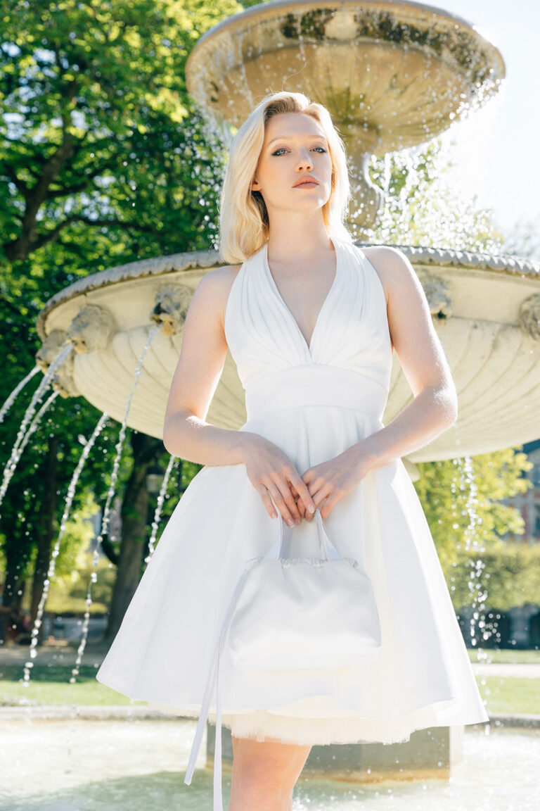 Femme habillée d'une robe de mariée courte sans manche qui tient un sac à main, devant une fontaine.