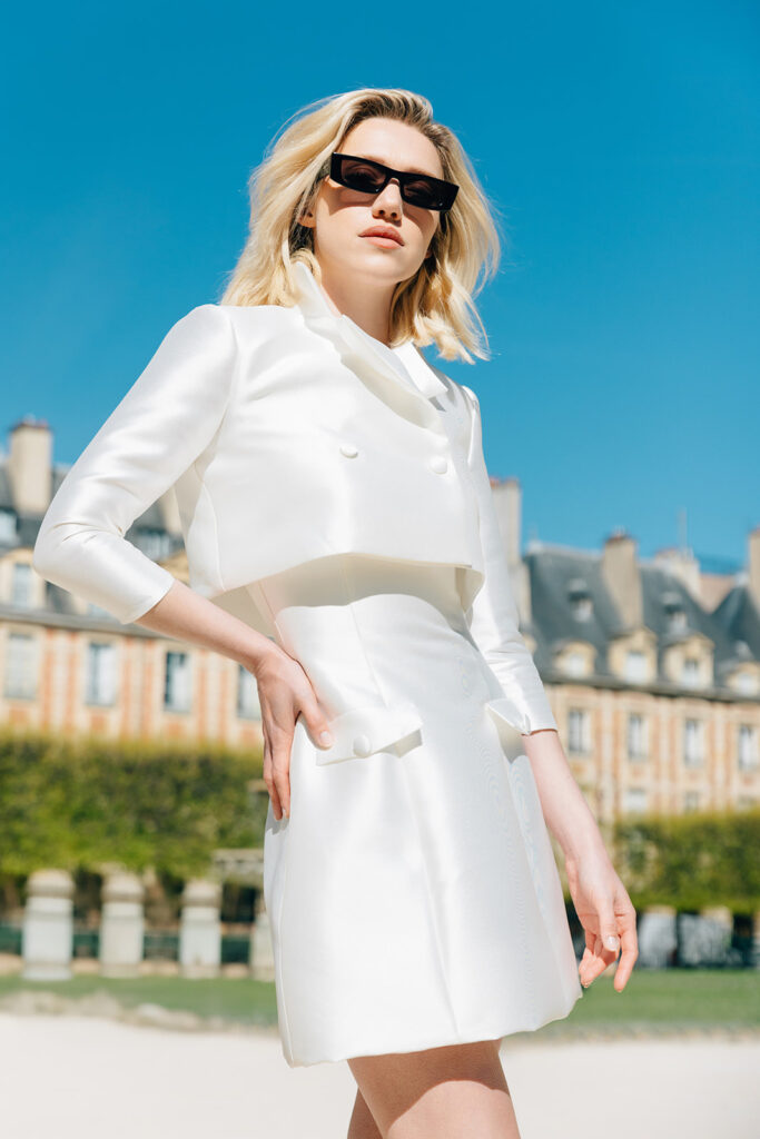 Femme habillée d'une robe de mariée courte accompagnée d'une veste blanche, et de lunettes de soleil.