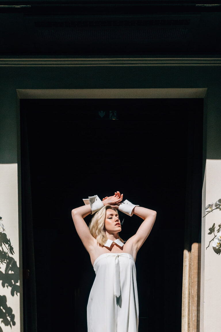 Femme qui lève les bras et qui porte une combinaison de mariée ample avec une cravate et les poignets et le col d'inspiration chemise.
