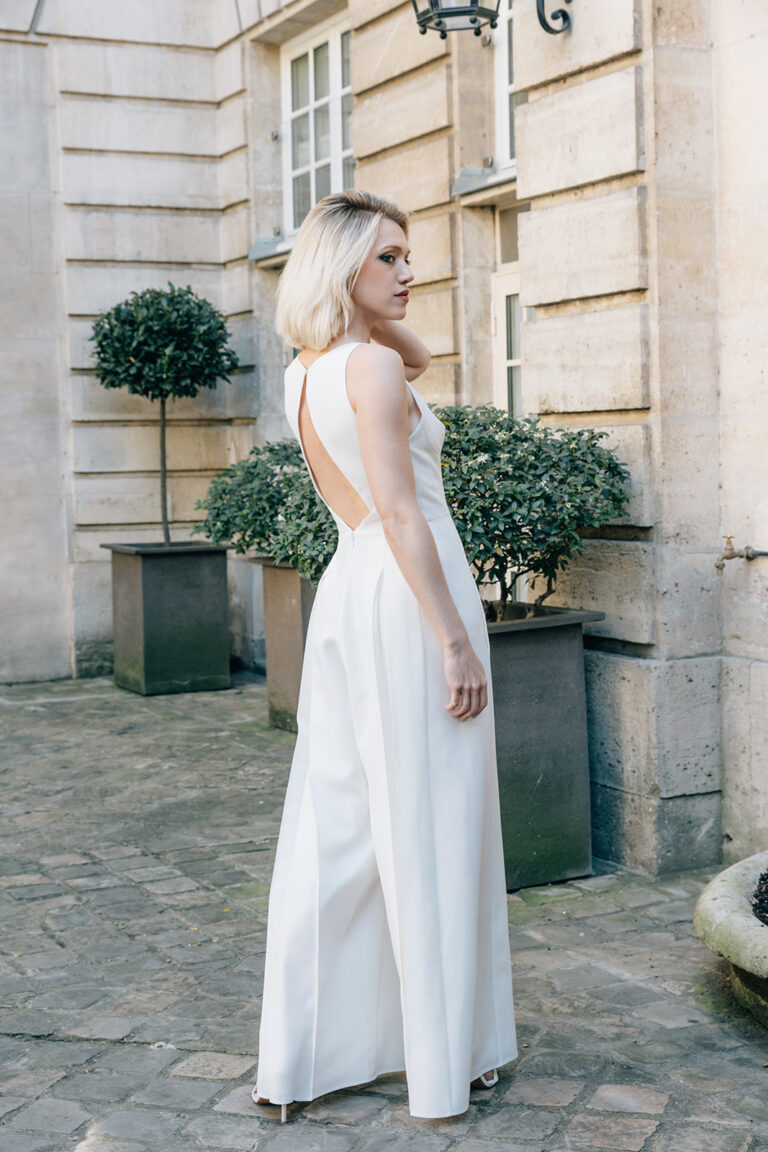 Femme qui est de dos et qui est vêtue d'une combinaison de mariée blanche et élégante, avec un joli dos nu.