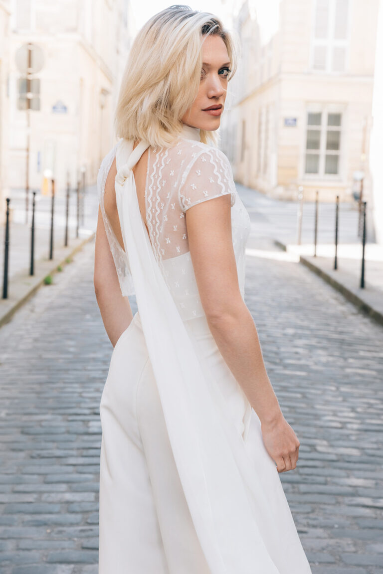 Femme portant une tenue de mariée blanche avec un pantalon et un top en dentelle de Calais-Caudry avec deux pants de tissus retombants dans le dos.
