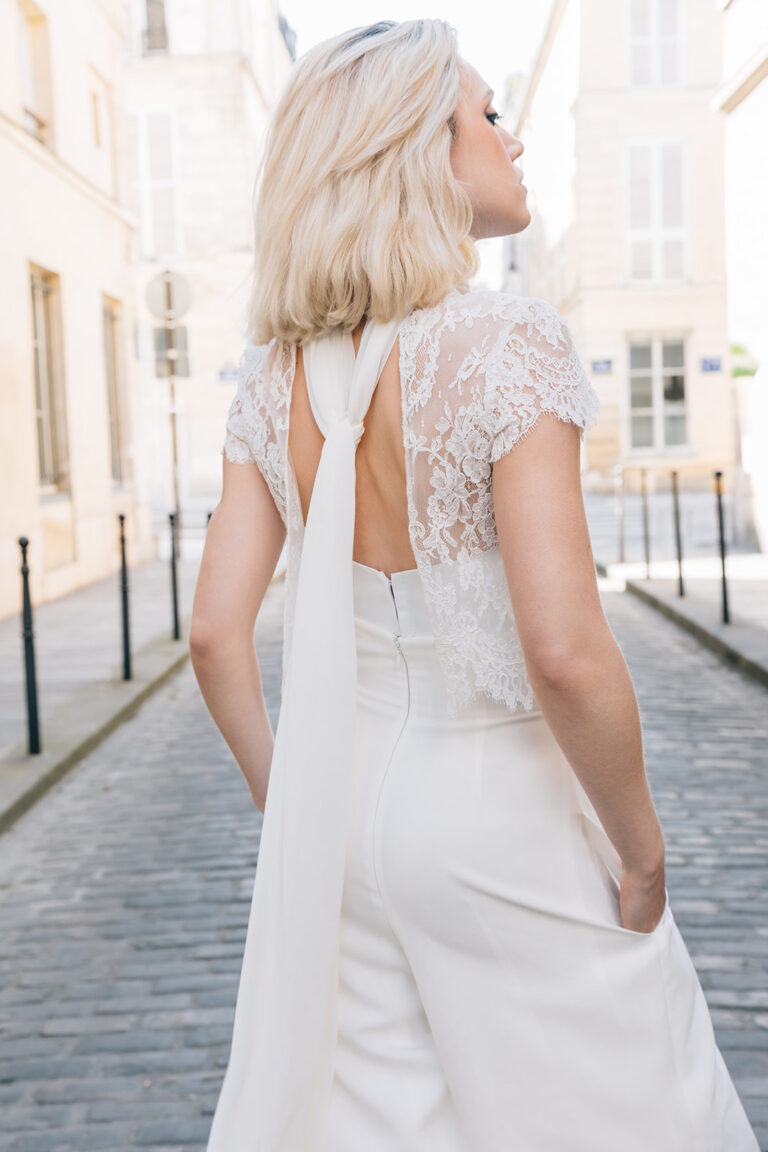 Femme portant une tenue de mariée blanche avec un pantalon et un top en dentelle de Calais-Caudry avec deux pants retombants dans le dos.