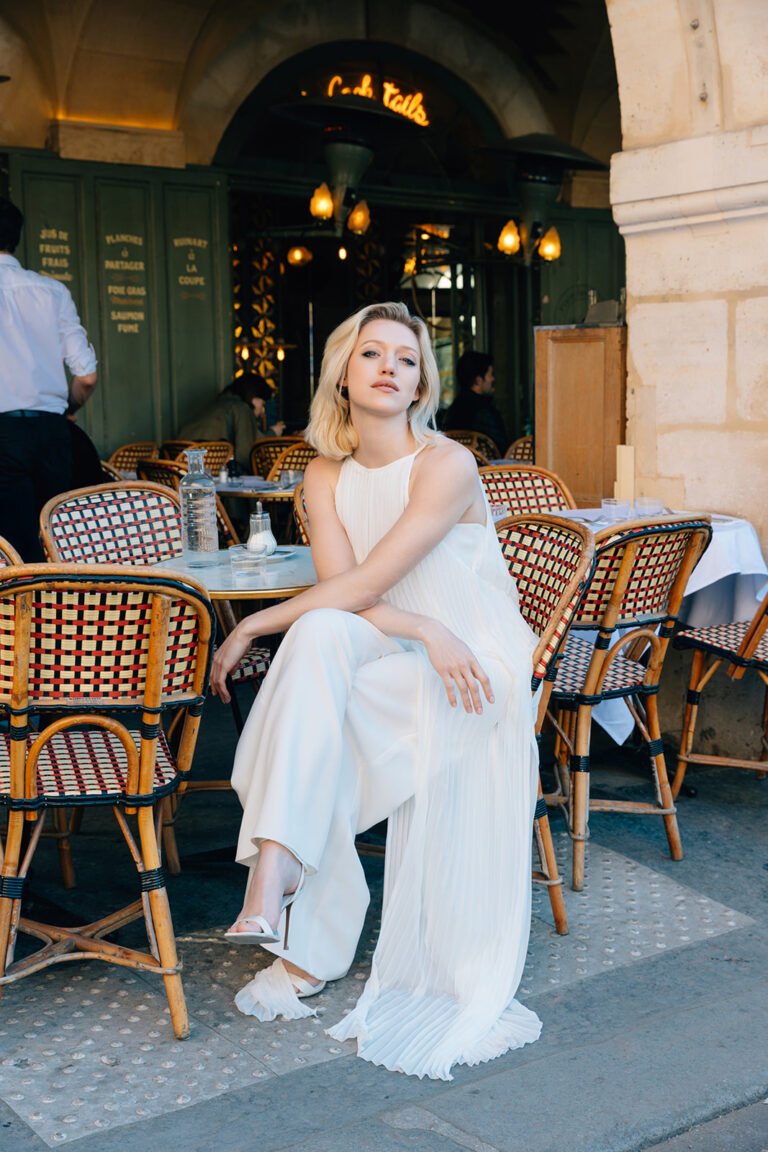 Femme assise à un café portant une tenue de mariée blanche avec un top long sans manches à mousseline plissé.