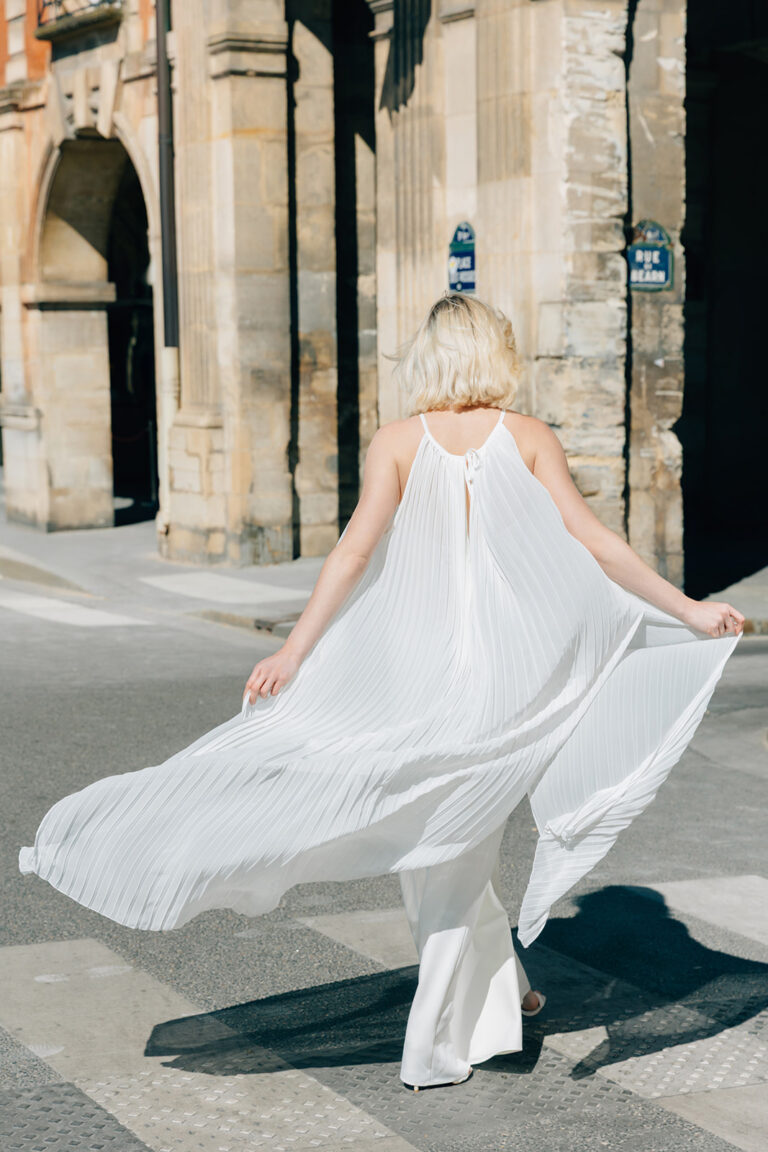 Femme de dos qui marche dans la rue en tenue de mariée blanche, avec un top long en mousseline plissé qui s'envole.