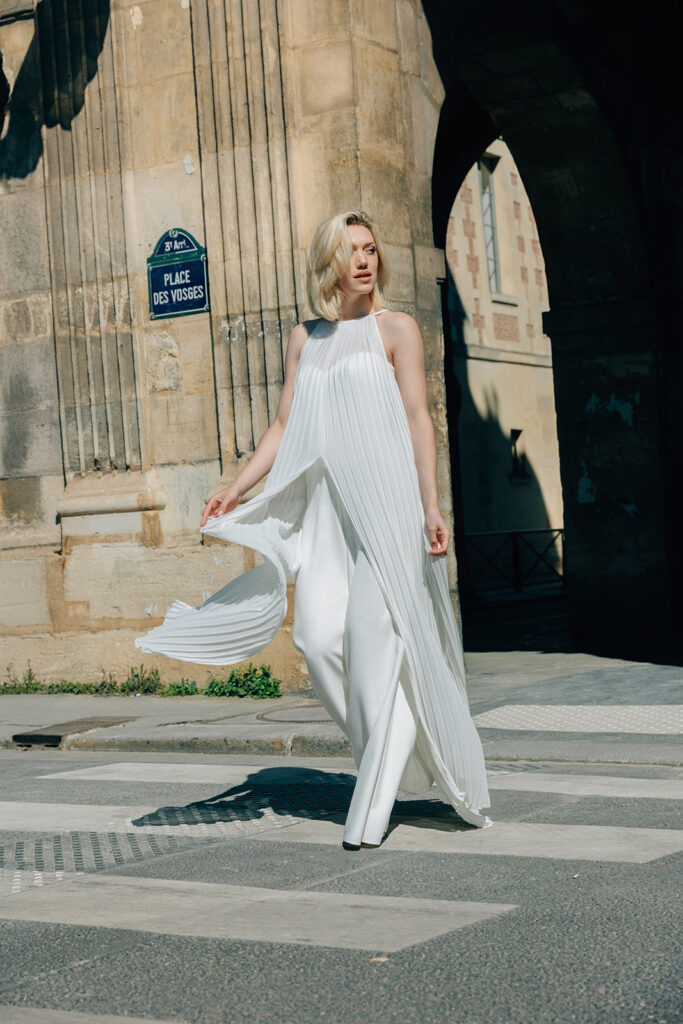 Femme qui marche dans la rue habillée d'une tenue de mariée blanche avec un top long sans manche à mousseline plissé.