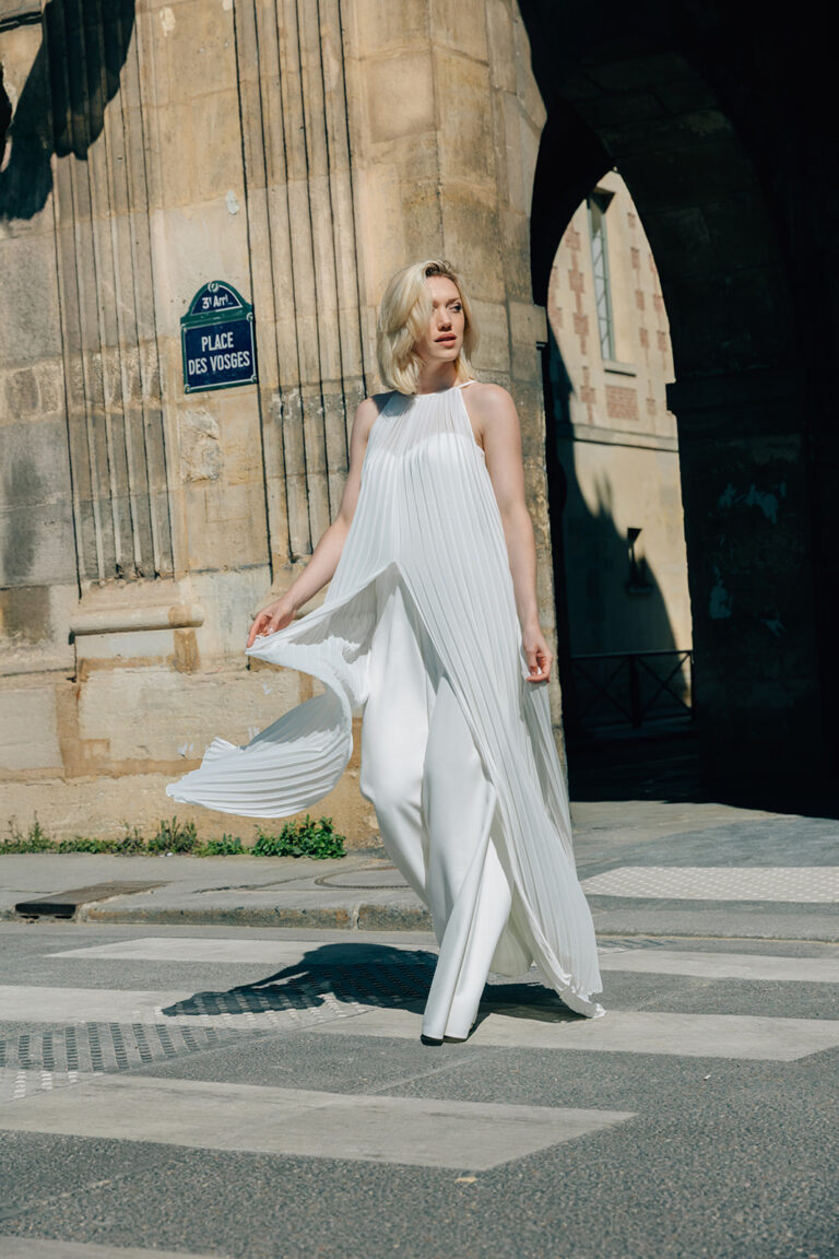 Femme qui marche dans la rue habillée d'une tenue de mariée blanche avec un top long sans manche à mousseline plissé.