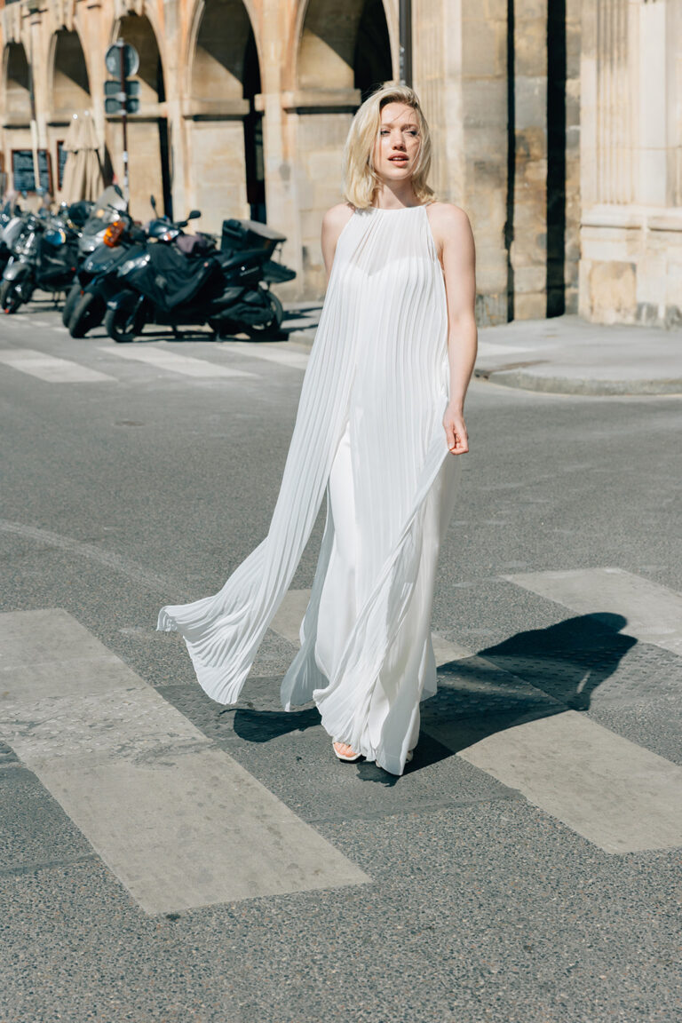 Femme qui marche dans la rue habillée d'une tenue de mariée blanche avec un top long sans manche à mousseline plissé.