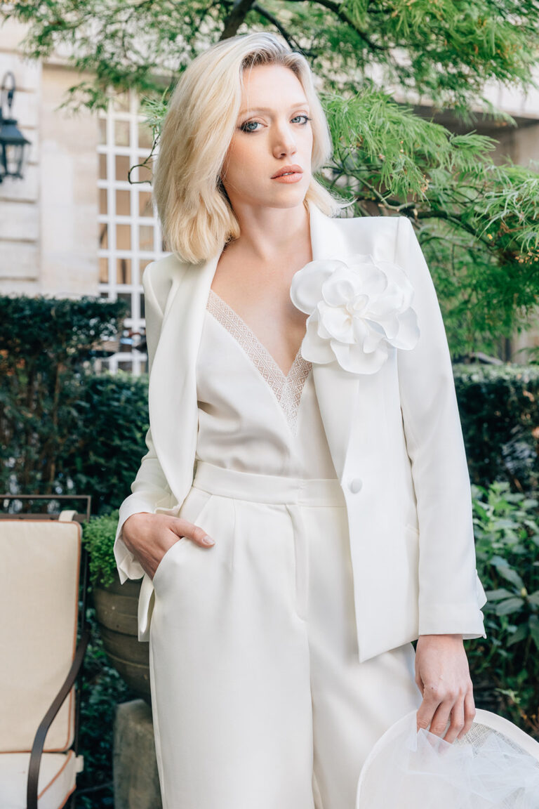 Femme qui porte un ensemble de mariée avec un pantalon, un top et une veste blanche avec une fleur.