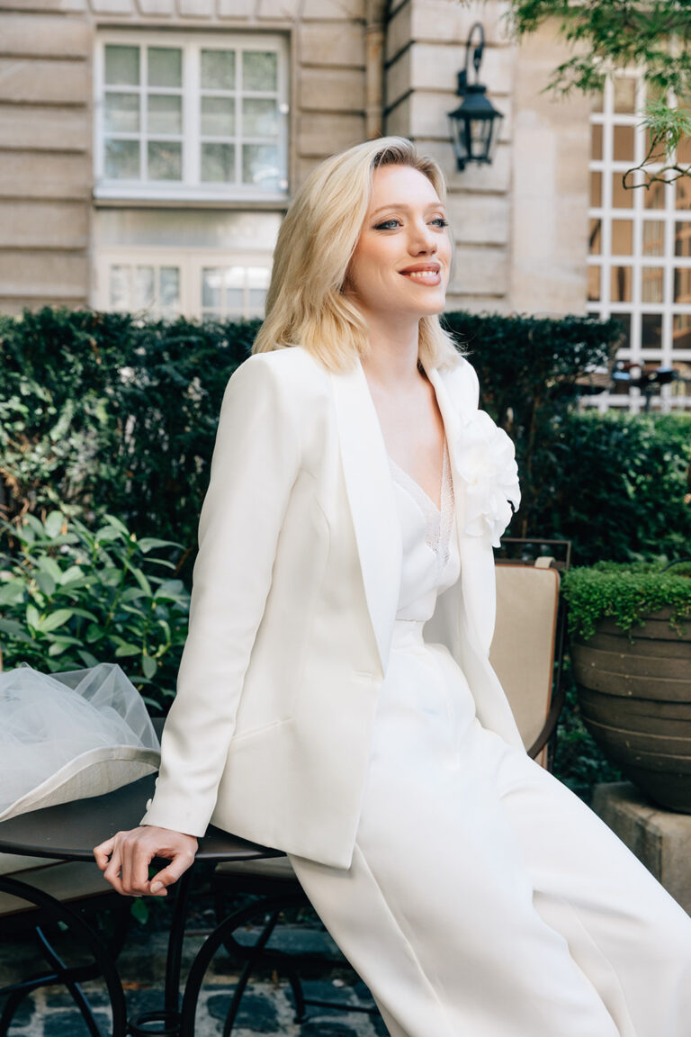 Femme assise qui porte un ensemble de mariée avec un pantalon, un top et une veste blanche avec une fleur.